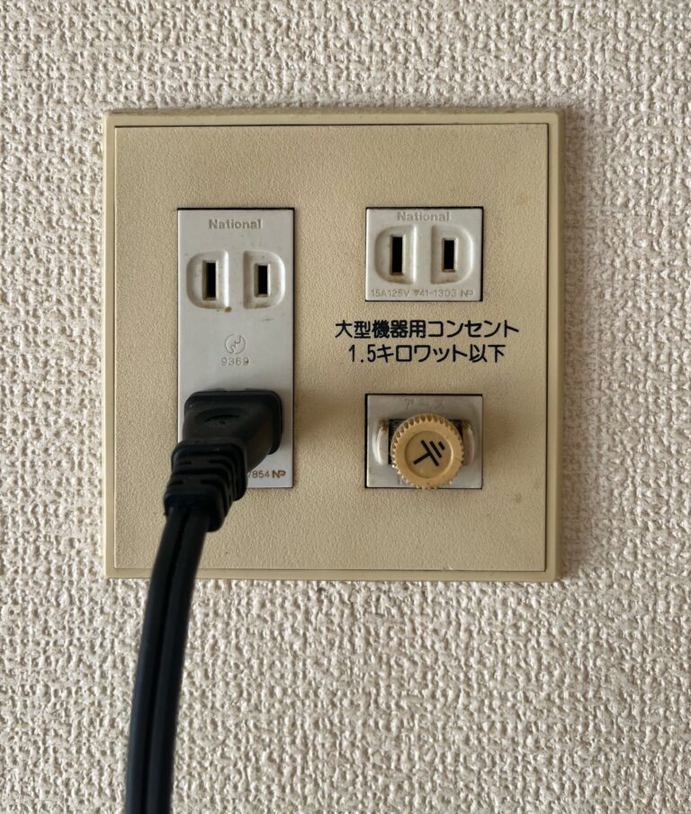 Beige topcontact in Japan met links twee stopcontacten onder elkaar. De onderste heeft een zwarte kabel erin. En rechtsboven nog een stopcontact met 2 gaten en daaronder een zekering. Het maakt eigenlijk twee gezichtjes.