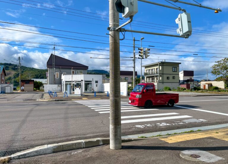 Een rood kleine auto met laadbak staat te wachten bij het stoplicht in het dorp. Niets is zo fel  en glimmend om 'm heen. 