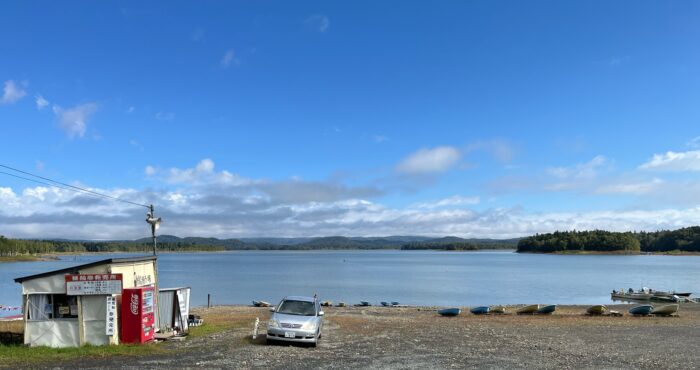Uitzicht op Lake Shumarina met daarachter bergen en bos. Op de voorgrond roeibootjes en een vervrommeld hokje en een enkele auto.