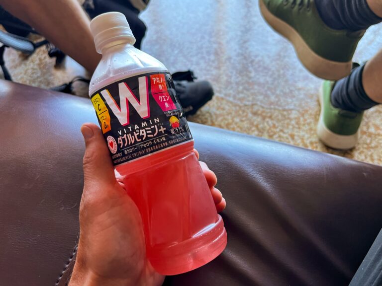 Een plastic flesje met een roode vloeistof erin met een zwarte wikkel met veel japans schrift en het woord vitamin. 