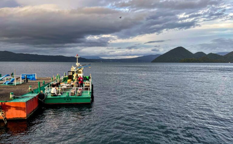 kleurrijke boot aan de steiger. Er zijn mannen bezig vuurwerk voor te bereiden. Op de achtergrond de eilanden van Lake Toya en een lucht vol regenwolken.