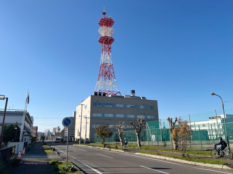 Boven op een vierkant grijs gebouw van 4 verdiepingen staat een metalen constructie. Een toren met horizontale witte en rode strepen. Het steekt mooi af tegen de heldere blauwe lucht.