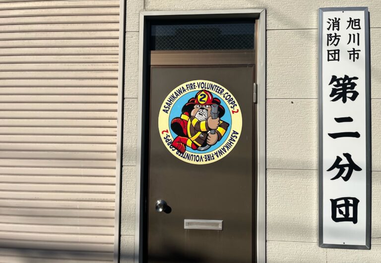 Een donkerbruine deur mat daarop een ronde stikker van een getekende buldog gekleed in een brandweerpak die een brandslang vasthoud. Daar omheen de tekst "Asahikawa fire volunteer corps 2"