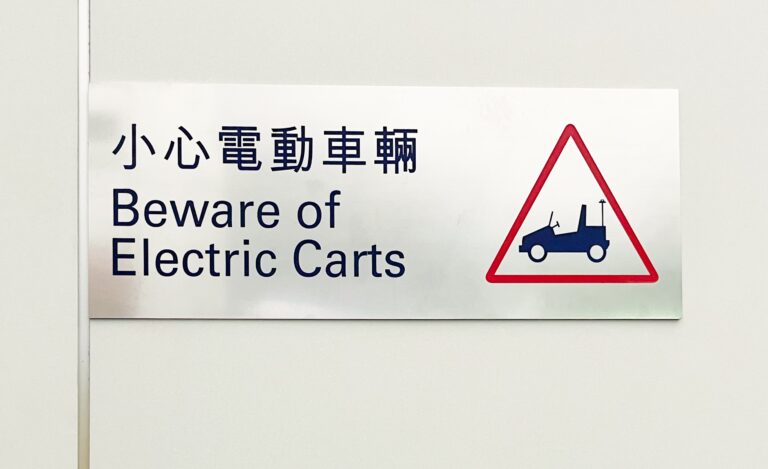 metalen bordje met daarop de waarschuwing "Beware of Electric Carts" Met daarnaast een pictogram van een elektrisch karretje in een rode driehoek.