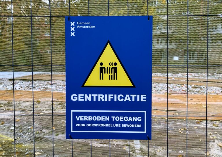 Blauw bord op een hek rond een gesloopt gebouw. Ziet er officieel uit als een bord van de gemeente Amsterdam. Met daarop een waarschuwingsdriehoek in zwart en geen met mensen erin. Daaronder: "Gentrificatie, verboden toegang voor oorspronkelijke bewoners"