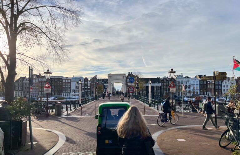 De magere brug in Amsterdam. Vanaf de oostkant net voor het oprijden met de fiets. Het is november en de zon staat laag en geeft een wat warme gloed de lucht is vol met kleine wolken als watten. Overal zijn natuurlijk fietsers.